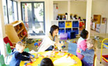 Children playing at table with teacher with floor play area in background.